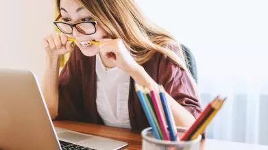 a long haired female student is sitting at a desk, staring intently at a laptop screen, chewing on a pencil.