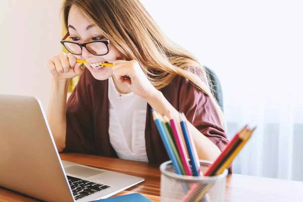a long haired female student is sitting at a desk, staring intently at a laptop screen, chewing on a pencil.